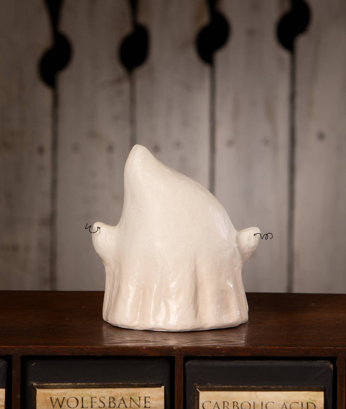 A ceramic ghost figurine in a glossy off-white glaze, displayed on a dark wooden surface with vintage apothecary-style drawers in the background.