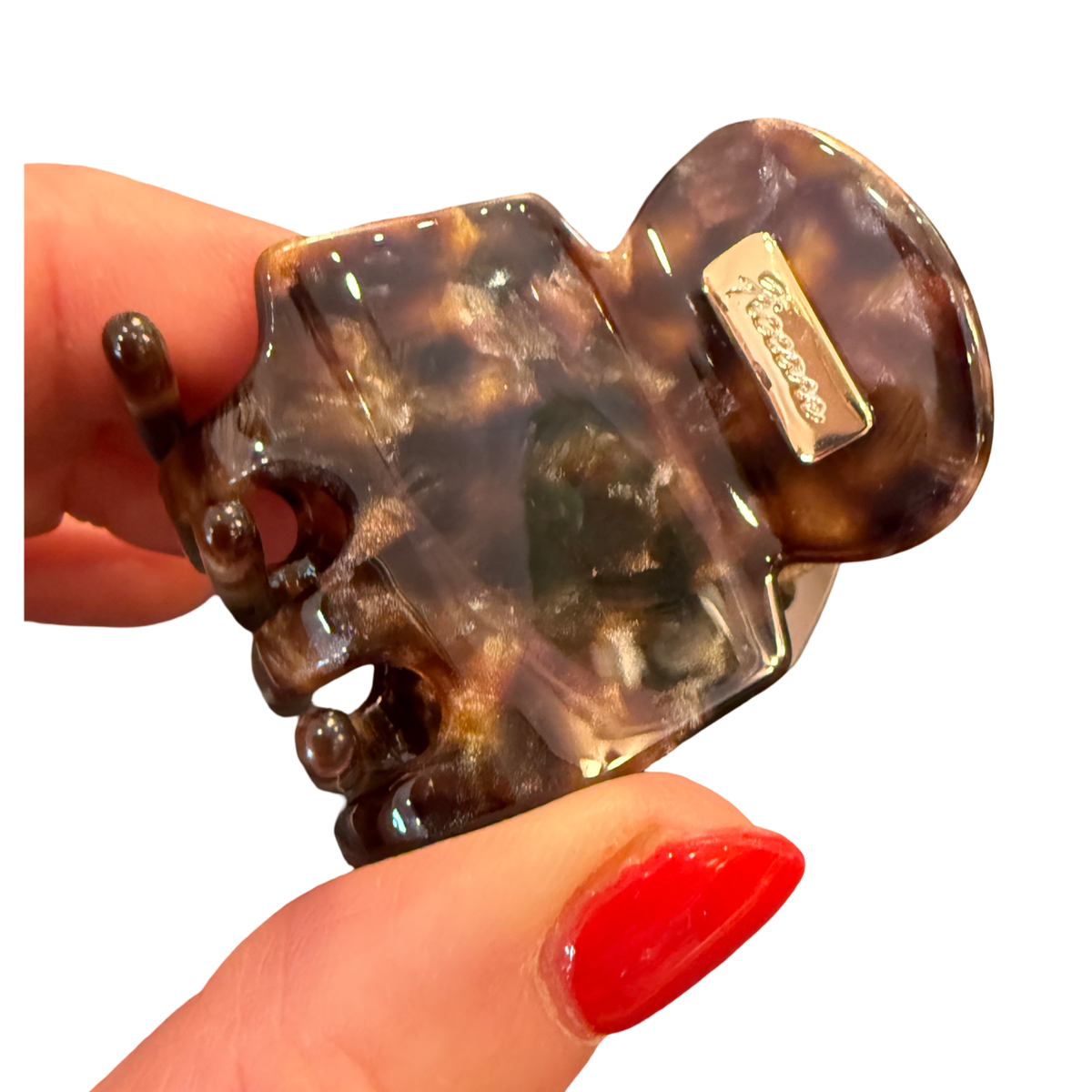 A tortoiseshell hair claw clip held by a hand with red nail polish against a plain white background. The brand name 'Sartoso' is visible on a small metal tag attached to the clip.