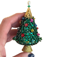 Hand-held close-up of a vintage green sequin Christmas tree ornament showing colorful pinned beads, an openwork gold-tone top, and a textured base.