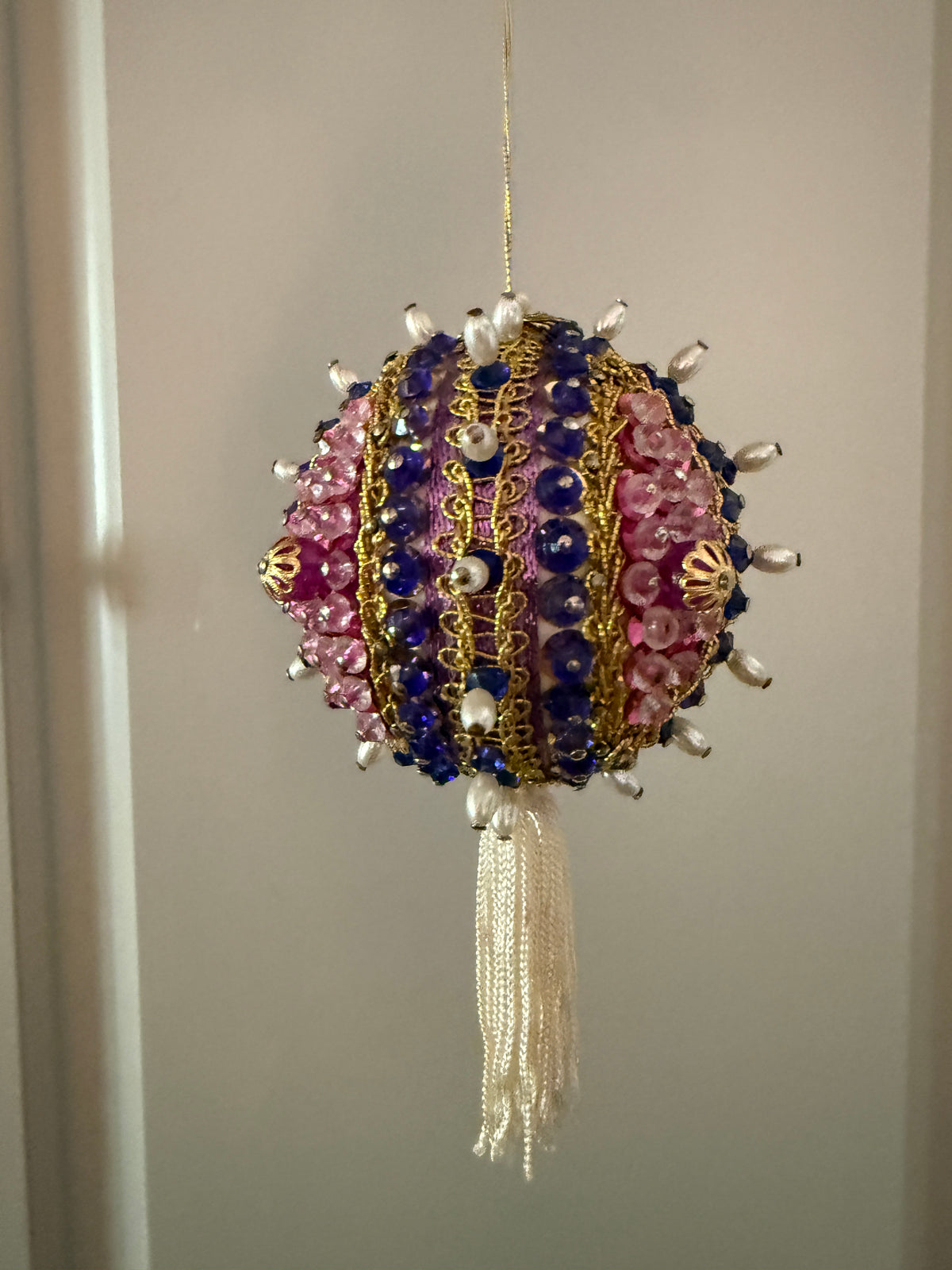 Close-up of a vintage purple and pink beaded ornament showing gold-tone floral details, decorative trim, pearl-look beads, and an ivory tassel.