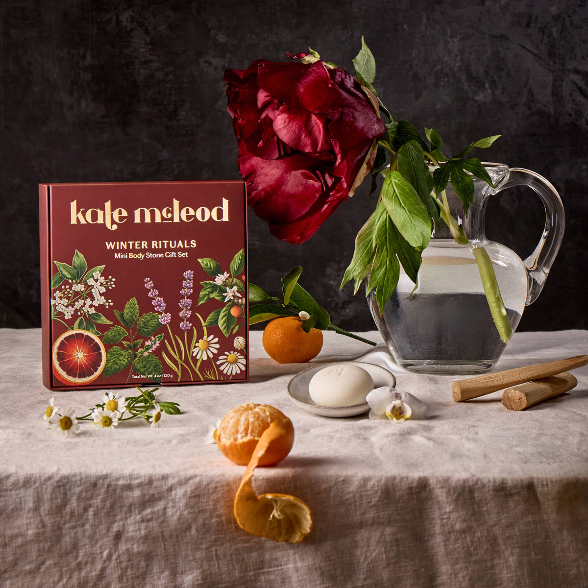 A boxed gift set labeled 'kate macleod winter rituals' sits on a draped fabric surface beside a glass pitcher, a large red rose, citrus, a white egg, small white flowers, and wooden sticks against a dark textured backdrop.