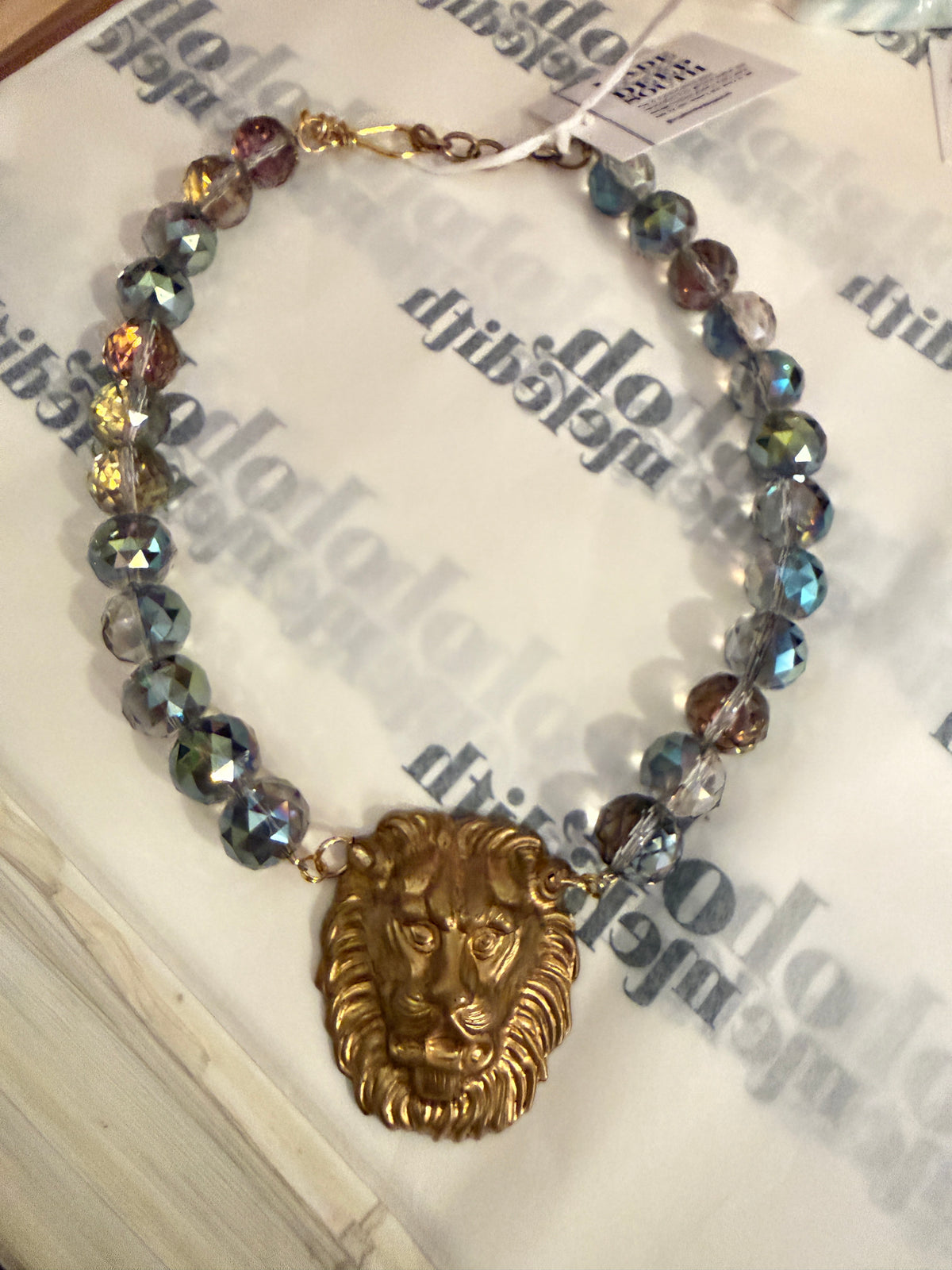 A beaded necklace with faceted iridescent beads in blue, green, and gold tones, featuring a prominent gold lion head pendant at the center, displayed on a printed white surface.