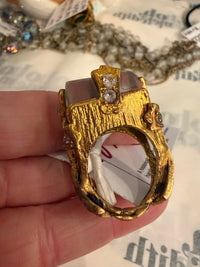 A close-up of a hand holding a chunky, organic-shaped gold ring with textured gold detailing, small clear rhinestones, and a translucent smoky gray stone.