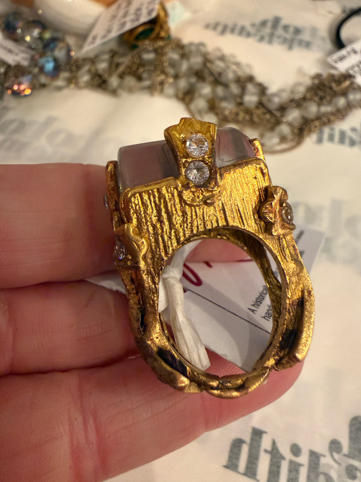 A close-up of a hand holding a chunky, organic-shaped gold ring with textured gold detailing, small clear rhinestones, and a translucent smoky gray stone.