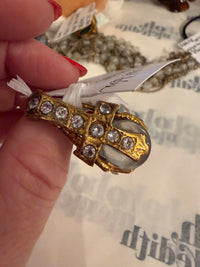 A gold ring with clear round gemstones and a large oval clear stone, held by a hand with red nail polish over a white background with faint text.