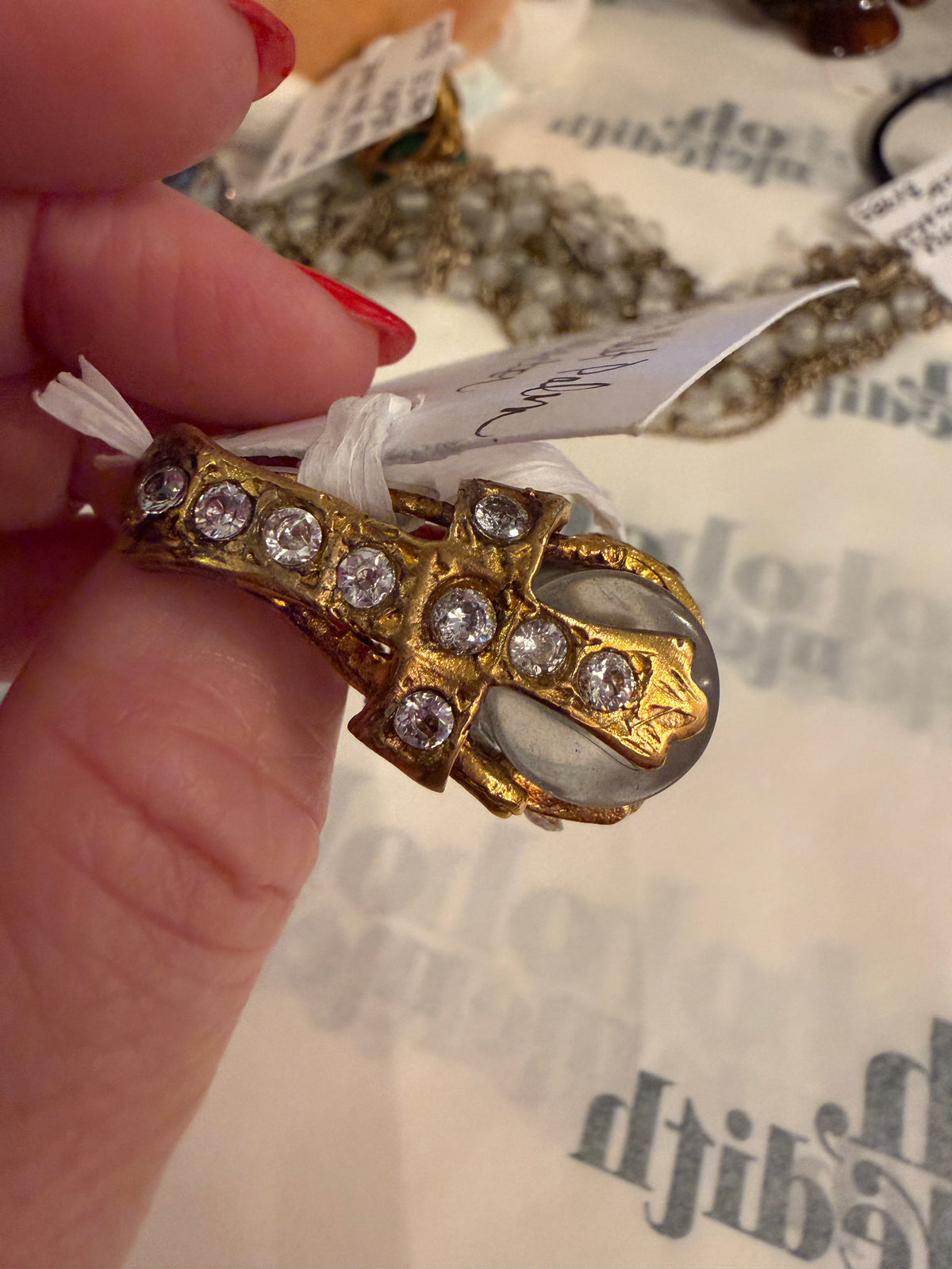 A gold ring with clear round gemstones and a large oval clear stone, held by a hand with red nail polish over a white background with faint text.