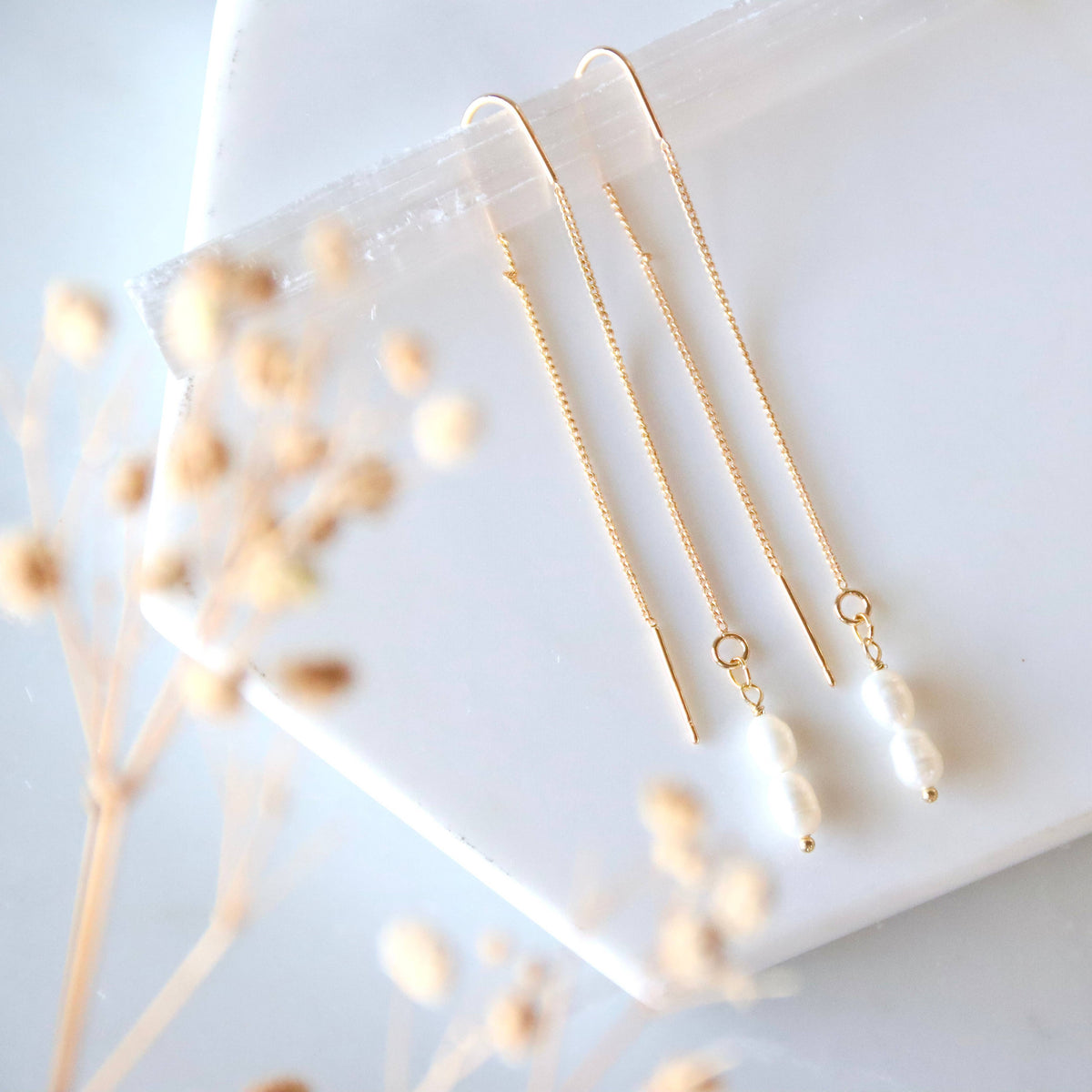 Two gold necklaces on a white surface with dried flowers.