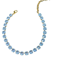 A gold-tone tennis necklace featuring light blue round-cut crystals, displayed on a clean white background.
