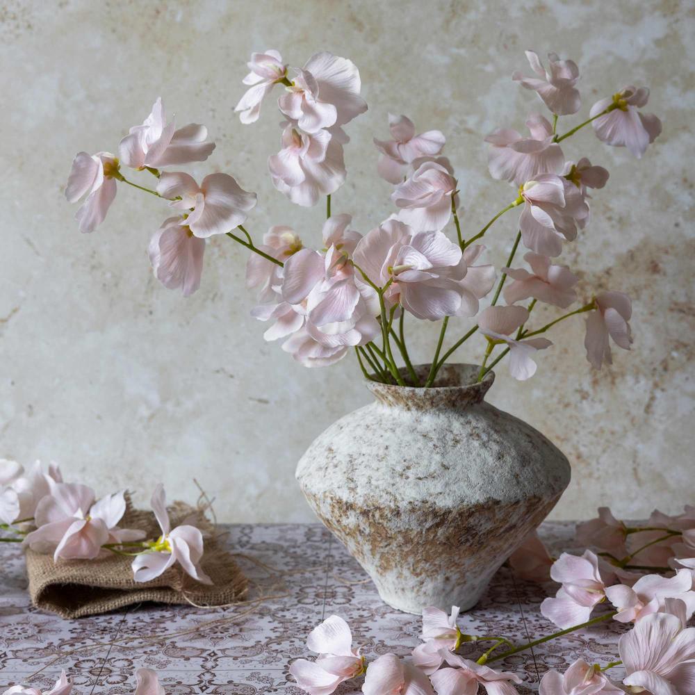 Pink flowers in a textured vase on a lace tablecloth with a floral background