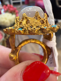 A close-up of a hand with red nail polish holding a gold ring featuring a crown-like top, a smooth white dome stone, and small turquoise blue accents.