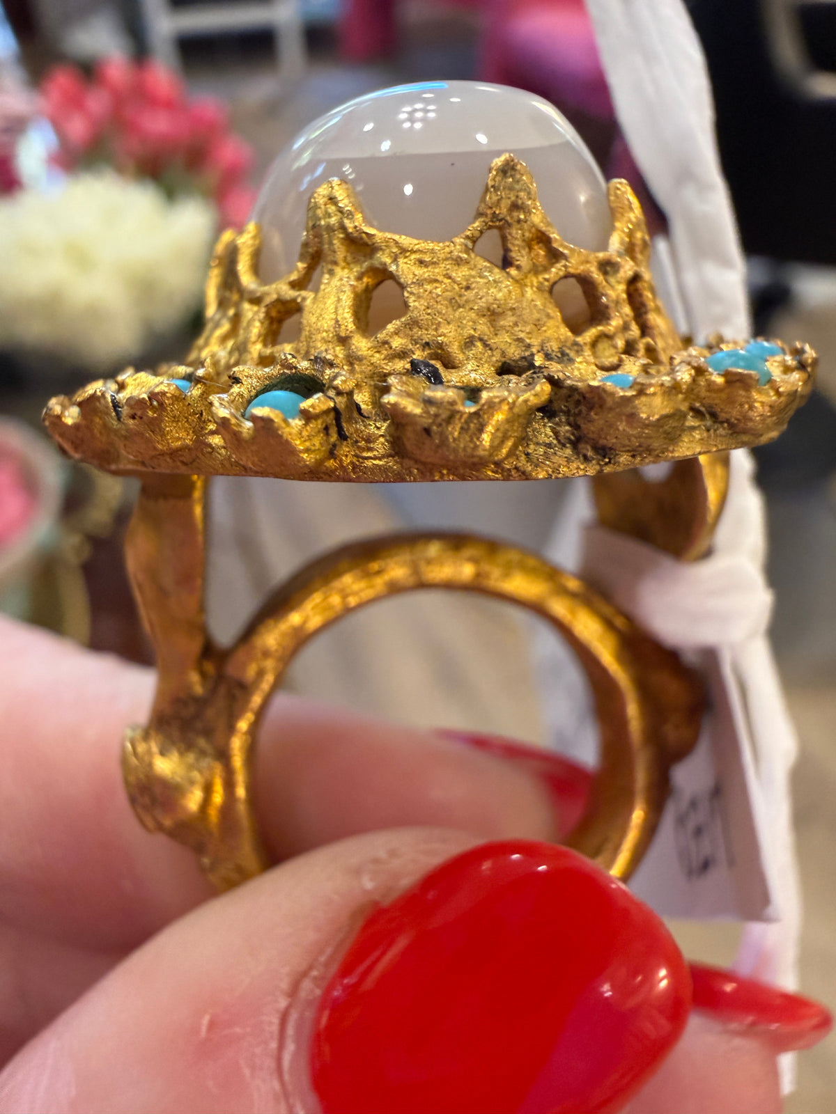 A close-up of a hand with red nail polish holding a gold ring featuring a crown-like top, a smooth white dome stone, and small turquoise blue accents.