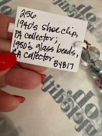 A close-up of a hand with red nail polish holding a white handwritten tag attached to a beaded shoe clip. The tag reads '256 1940's shoe clip, PA collector, 1950's glass beads, PA collector 81817'.