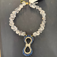 A beaded necklace with faceted clear crystal-like beads and a central infinity-shaped pendant decorated with blue stones and gold-toned metal, shown on a dark gray surface with a branded tag attached.