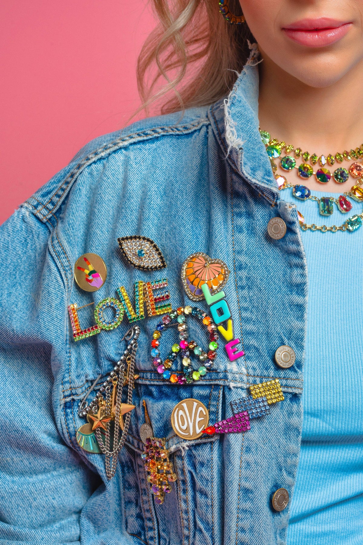 Person wearing a denim jacket with colorful patches and accessories against a pink background