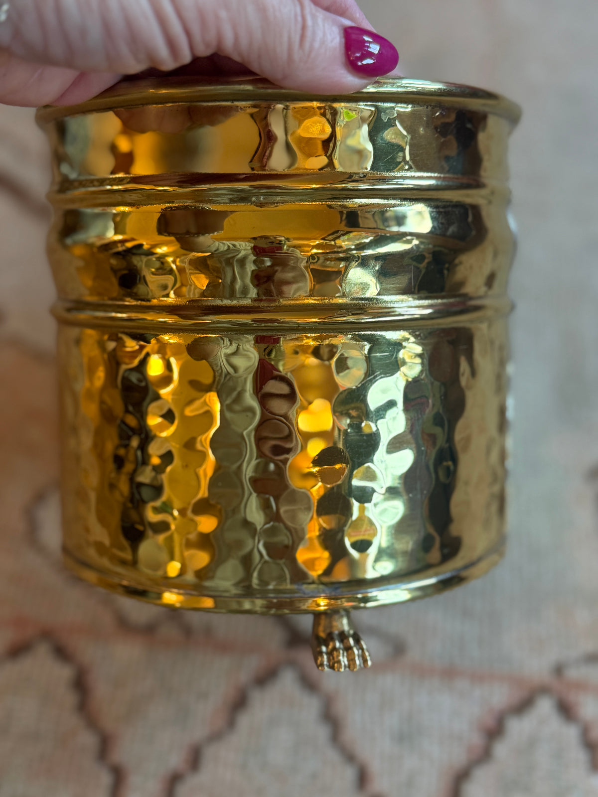 Gold textured container held by a hand with pink nail polish on a patterned background